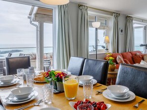 Una mesa de comedor dispuesta con alimentos para el desayuno, vista al mar y un rincón de estar acogedor con un sofá y una lámpara.