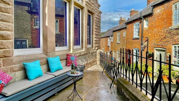A stone building with large windows, a stone bench with cushions, a small table with a glass, and a metal fence. - 1 Bed in Matlock (oc-86416) (Wirksworth)