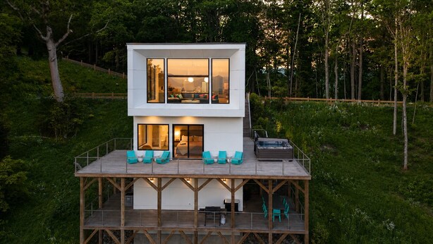 Una casa moderna de dos pisos con un balcón, mobiliario exterior y vistas al bosque circundante.