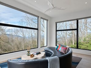 Una sala de estar moderna con un sofá curvo, una mesa de centro redonda y vistas a un paisaje boscoso a través de amplios ventanales.