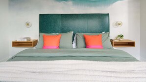 Una cama con cabecera verde, dos almohadas naranjas y una manta blanca y mullida, además de un mural en la pared.