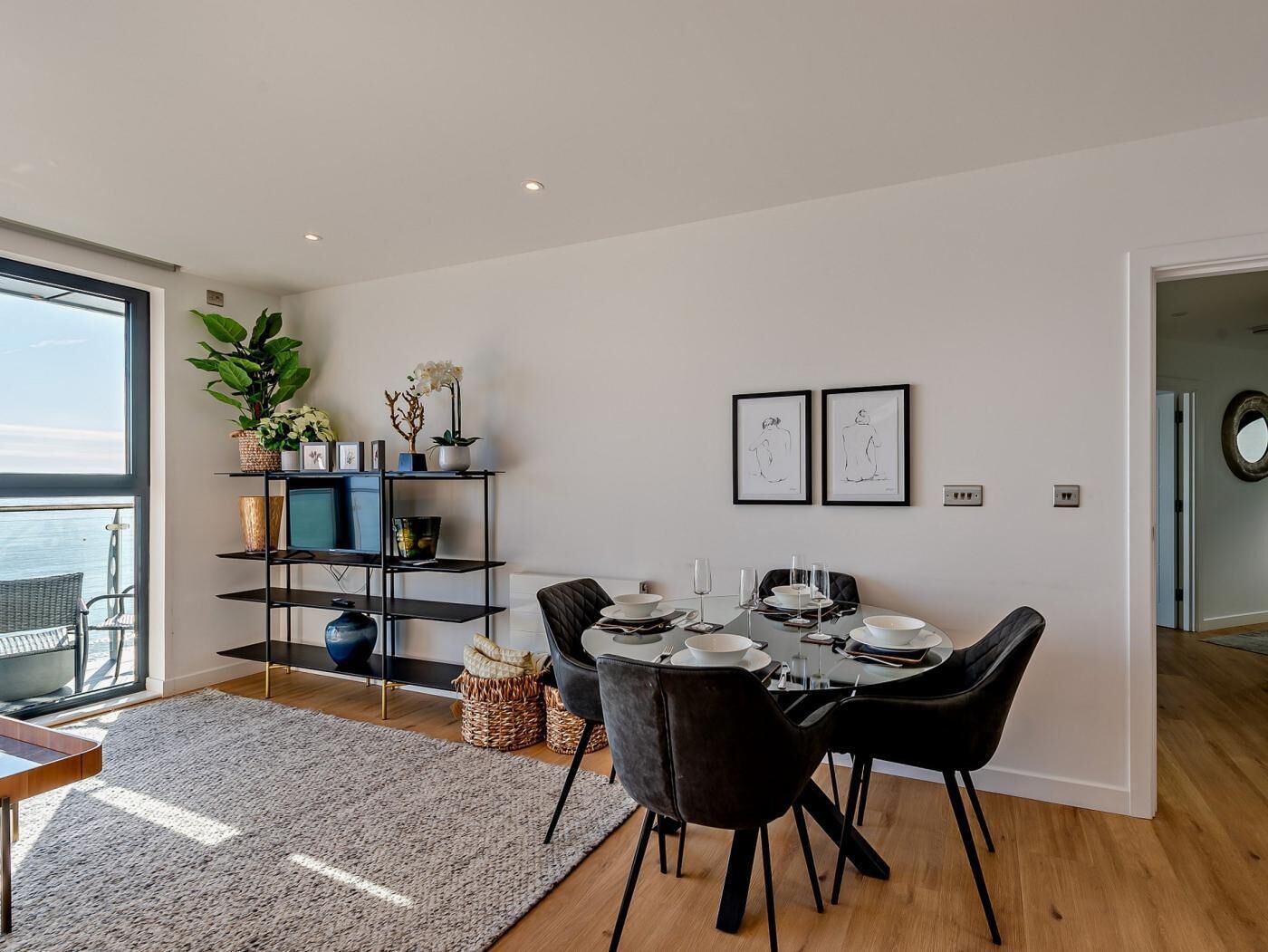 Un espace de repas moderne avec une table dressée pour quatre personnes, une chaise noire, une bibliothèque agrémentée d’objets décoratifs et une grande fenêtre offrant une vue.