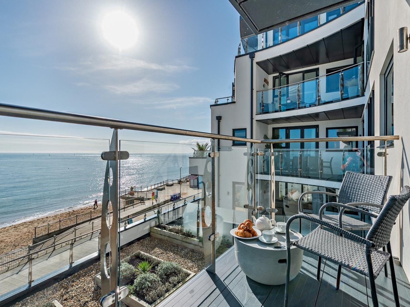 Un balcon avec vue sur la mer, une table ronde avec une théière et des pâtisseries, et une chaise.