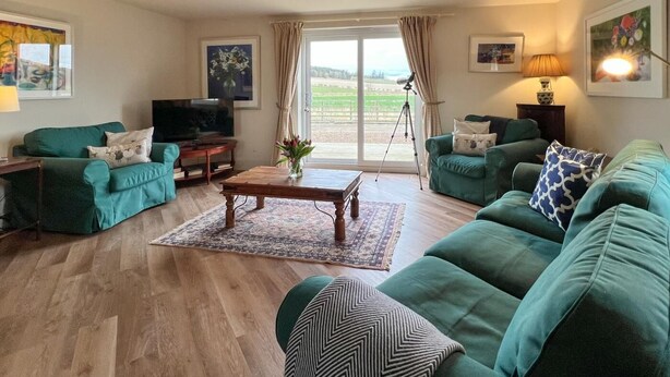 A living room with teal sofas, a wooden coffee table, a TV, and a large window with curtains. - 2 Bed in Mindrum (oc-86637) (Carham)