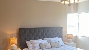 Ein Schlafzimmer mit einer gesteppten Kopfteile, einem Bett mit gestreifter Bettwäsche, zwei Nachttischen mit Lampen sowie einem Fenster mit Tageslicht.