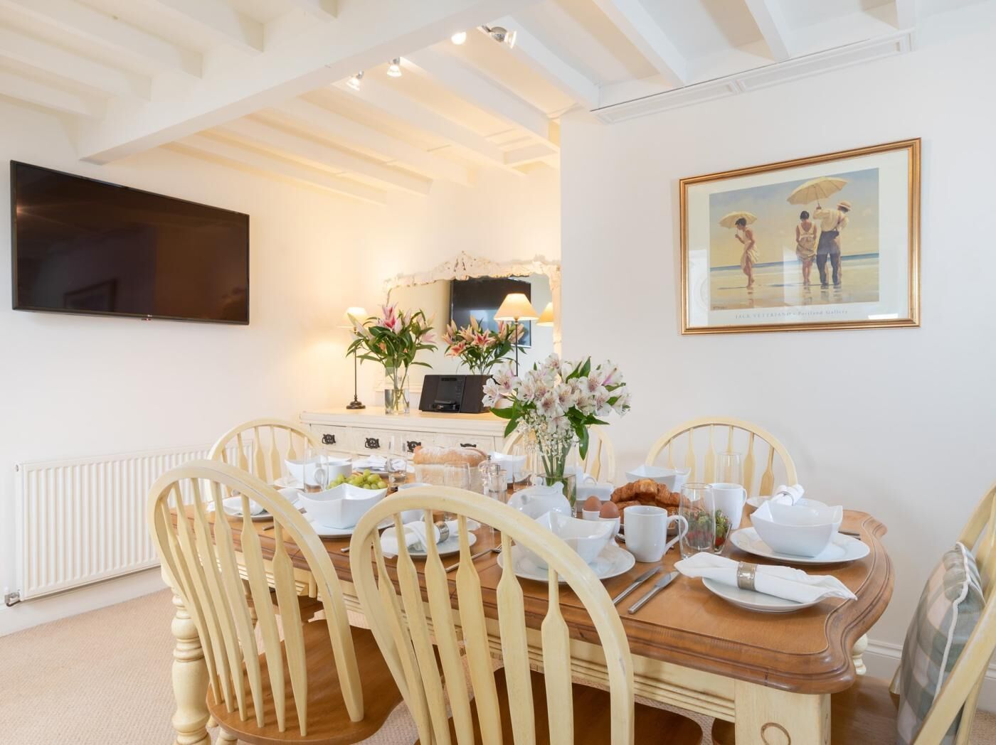 Un coin repas avec une table en bois dressée pour le petit-déjeuner, des chaises blanches, un radiateur blanc, un téléviseur à écran plat et un tableau encadré accroché au mur.