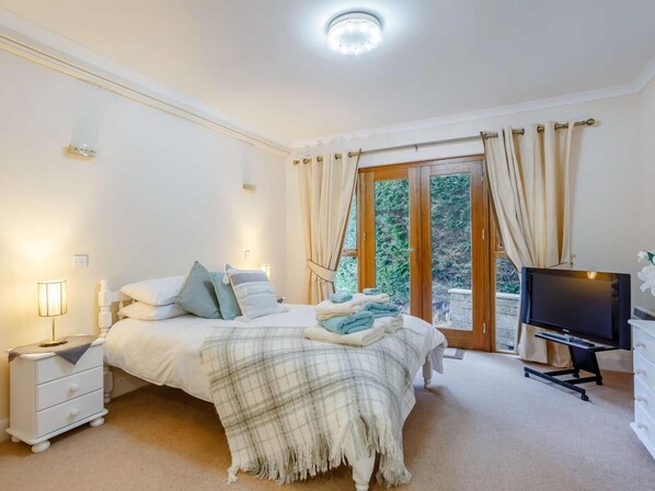 A bedroom with a bed, bedside table, TV, and a view of greenery through the door.