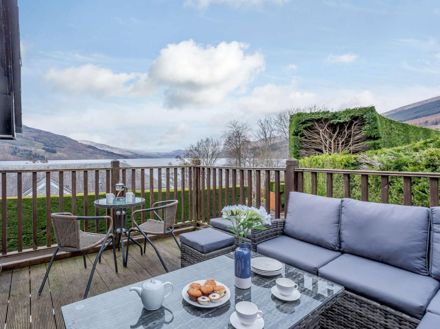 En balkon med udsigt over et sø, et bord med tekande og muffins, og en sofa med puder.