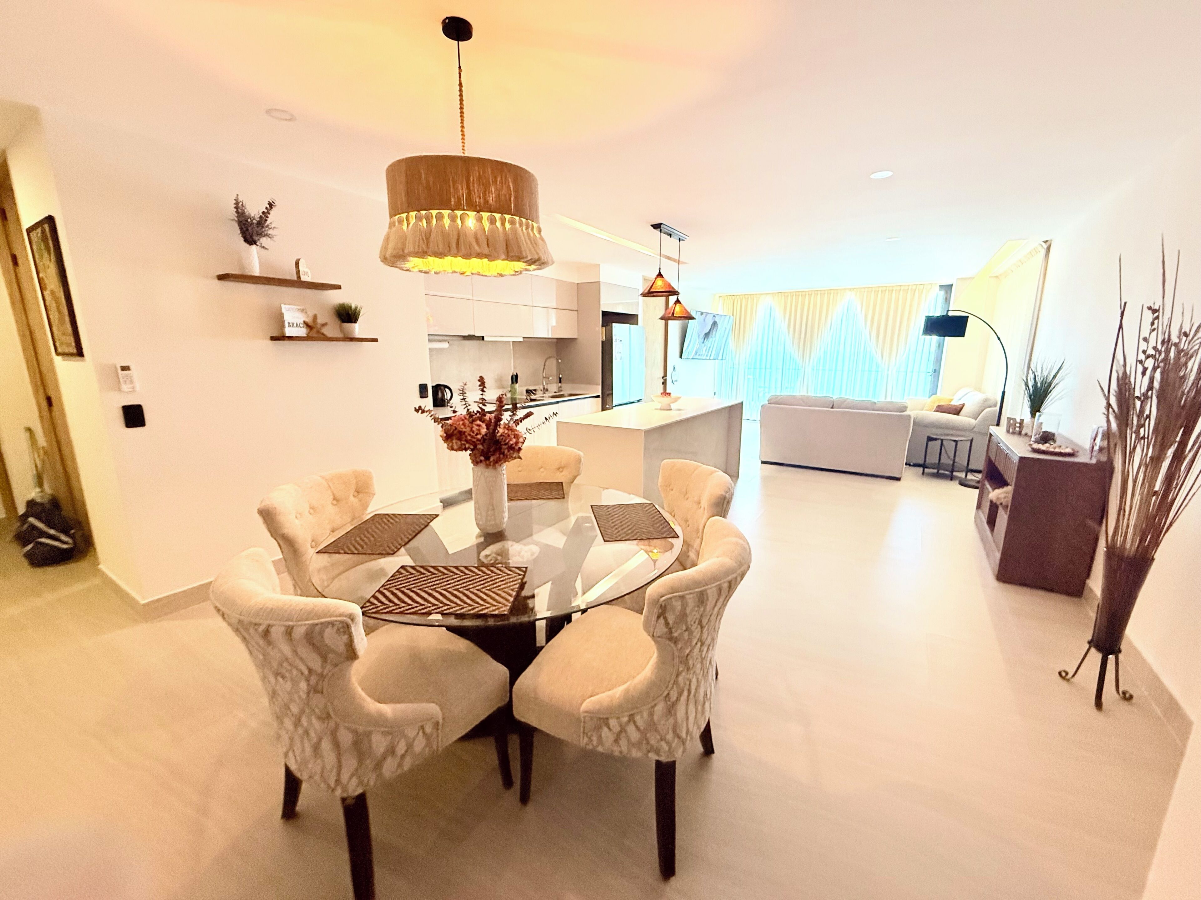 Ein modernes Wohnzimmer mit einem runden Glastisch, weißen Sesseln, einer Pendelleuchte und einer Küche im Hintergrund.