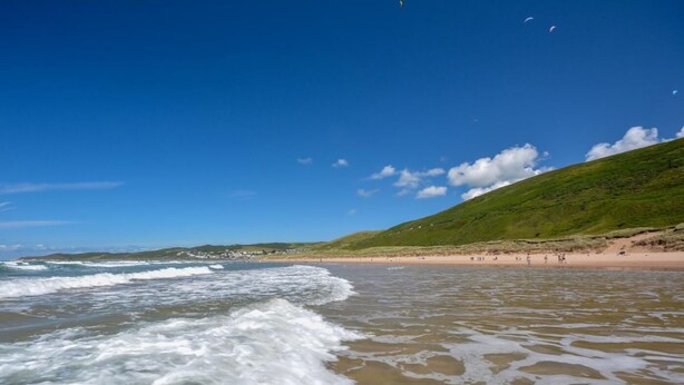 En strand med vågor, en sandstrand och en kulle med grönskande vegetation.