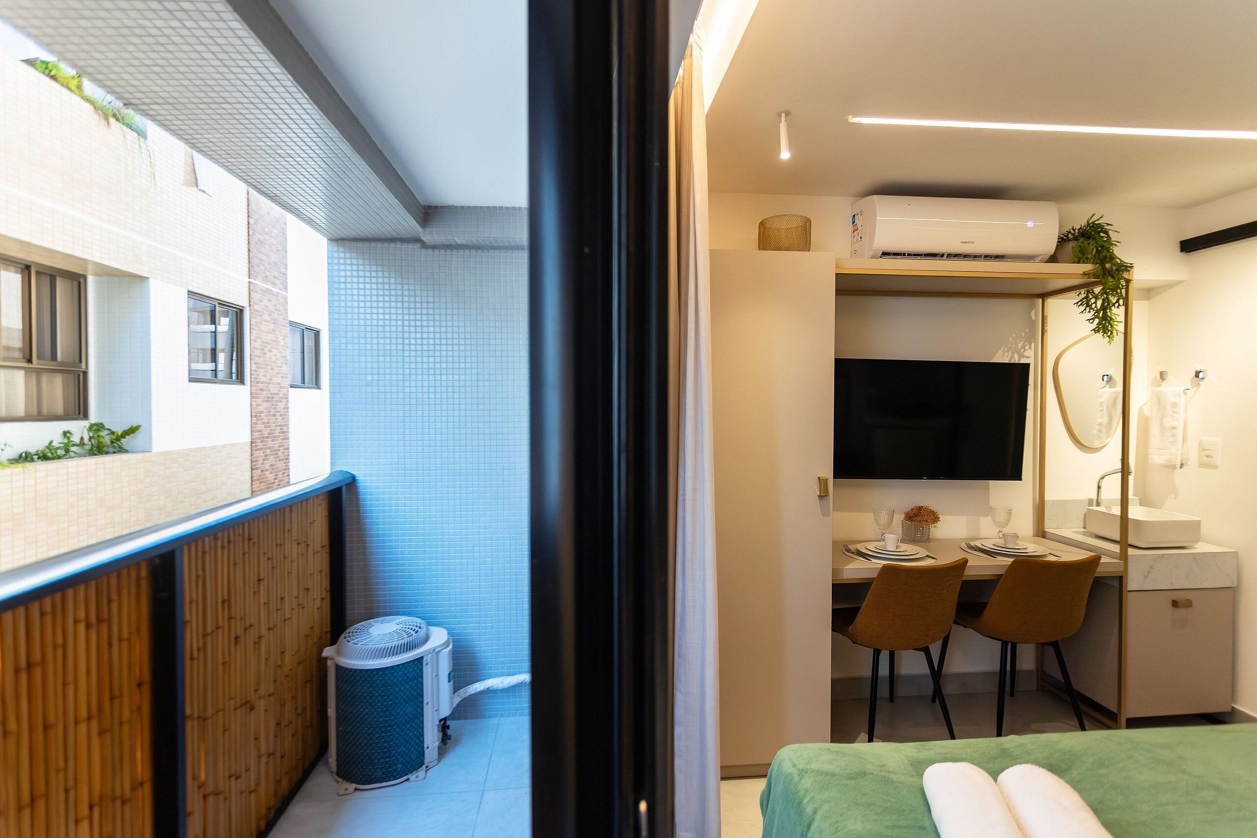 A compact hotel room with a balcony, a small desk with chairs, a television, and a bed with a green bedspread.