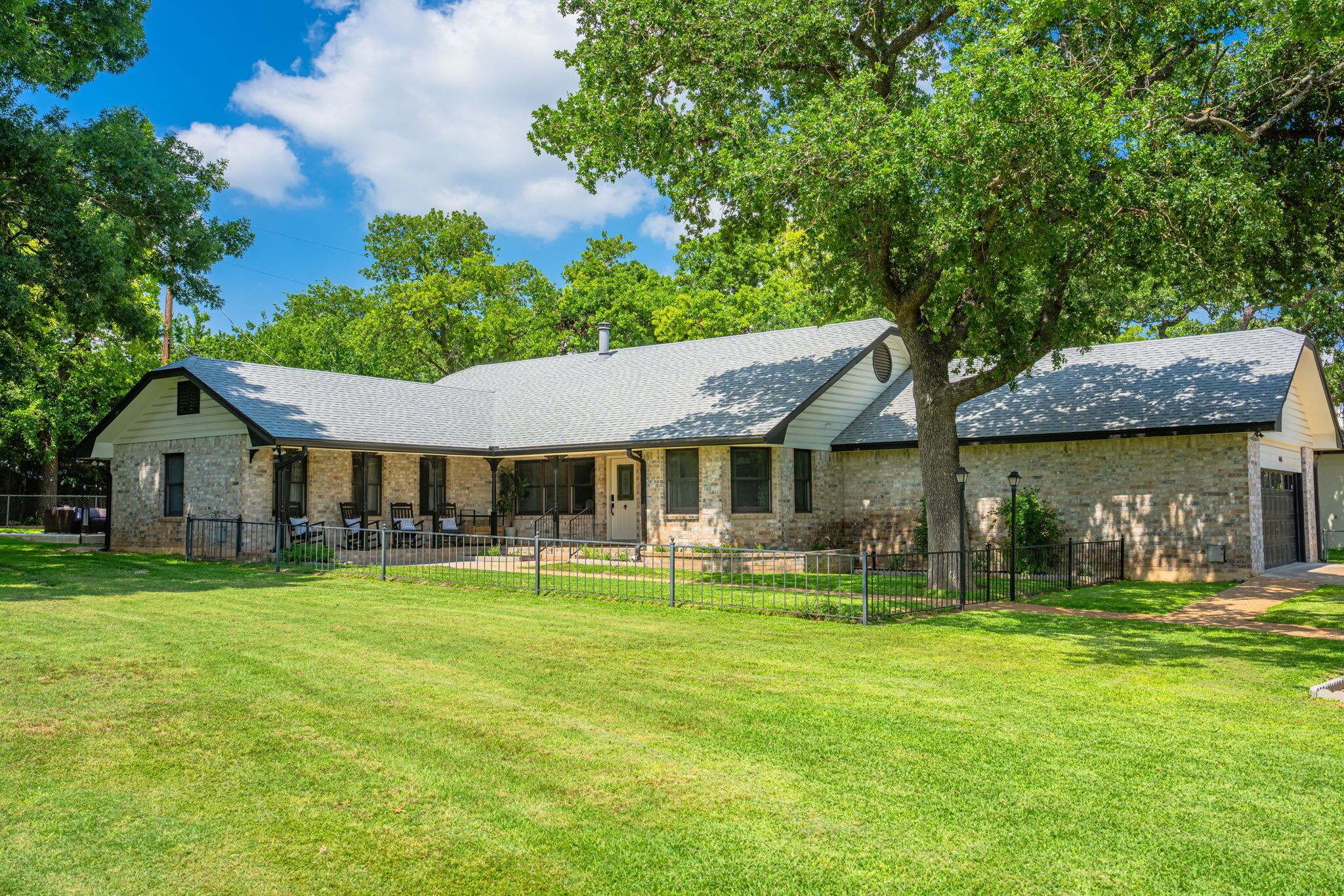 A two-story stone building with a covered porch, a fenced yard, and trees in the background.
