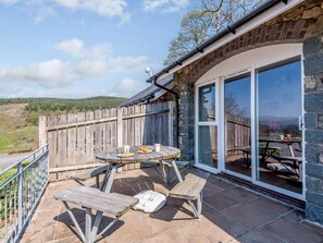 Eine Terrasse mit Tisch und Bänken, ein Holzzaun und ein Steinhaus mit Glastür.