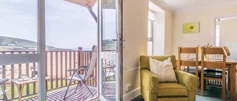 Una habitación con puerta corrediza de vidrio que da a una terraza exterior, un sillón verde, una mesa de comedor de madera y un cuadro en la pared.