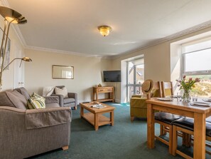 Ein Wohnzimmer mit Sofa, Couchtisch und Esstisch. Ein Fernseher ist an der Wand montiert und ein Fenster bietet einen Ausblick.