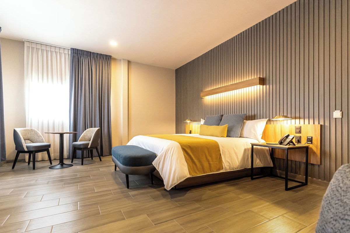 A modern hotel room with a bed, bedside tables, a chair, and a desk.