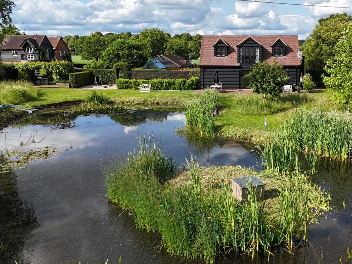 A serene pond with surrounding greenery and a traditional house with a pitched roof.