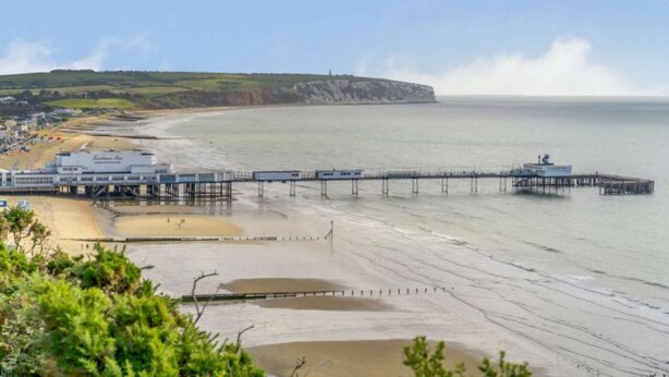 A coastal area with a pier extending into the sea, a beach, and a cliff in the background. - 5 Bed in Sandown (oc-87008) (Sandown)