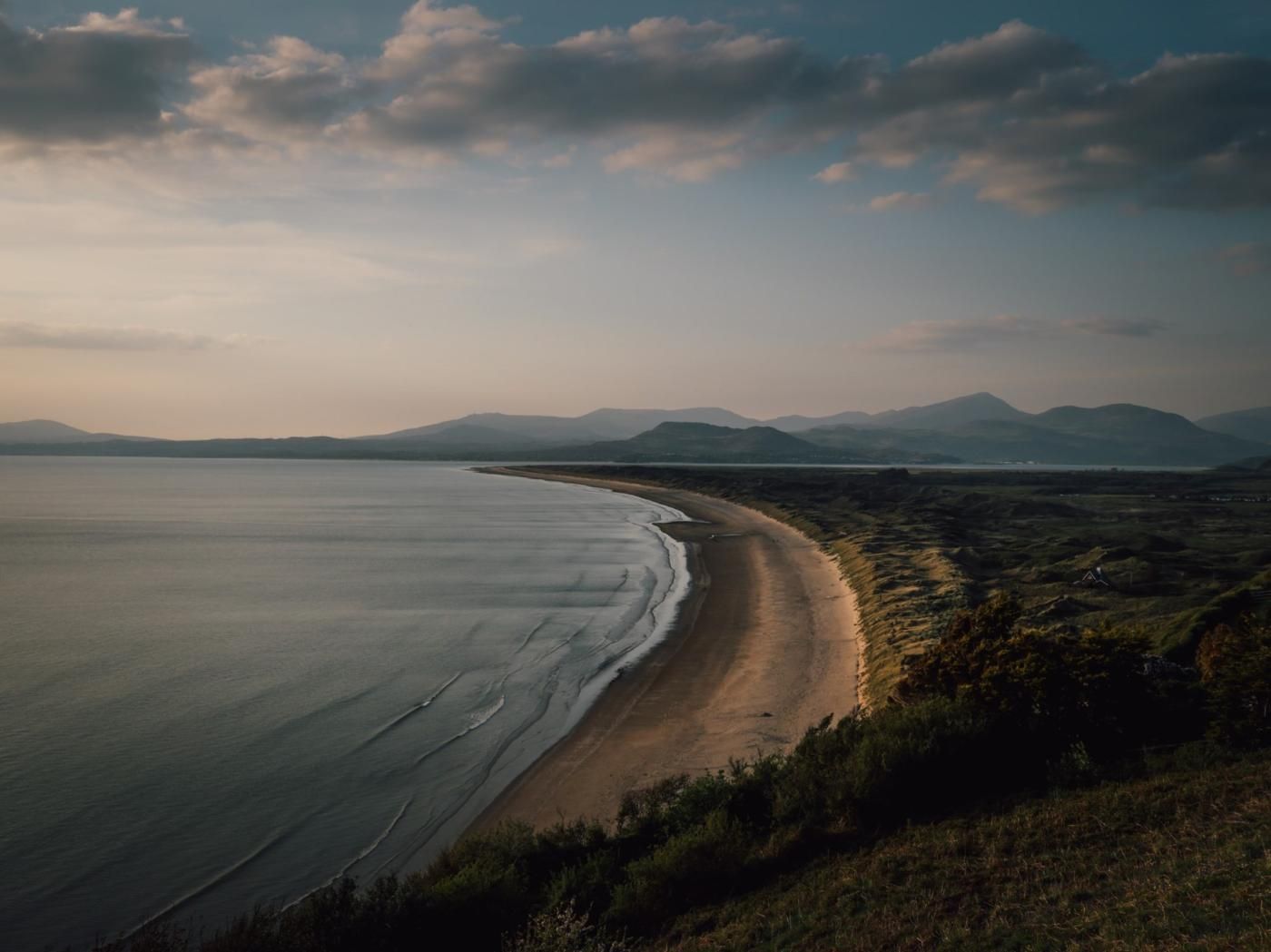 Een kustlandschap met een zandstrand, grasvelden en verafgelegen bergen, alles onder een bewolkte lucht.
