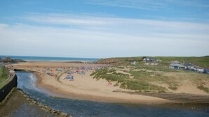 Une plage côtière bordée d’une rivière, de dunes herbeuses et de quelques bâtiments.
