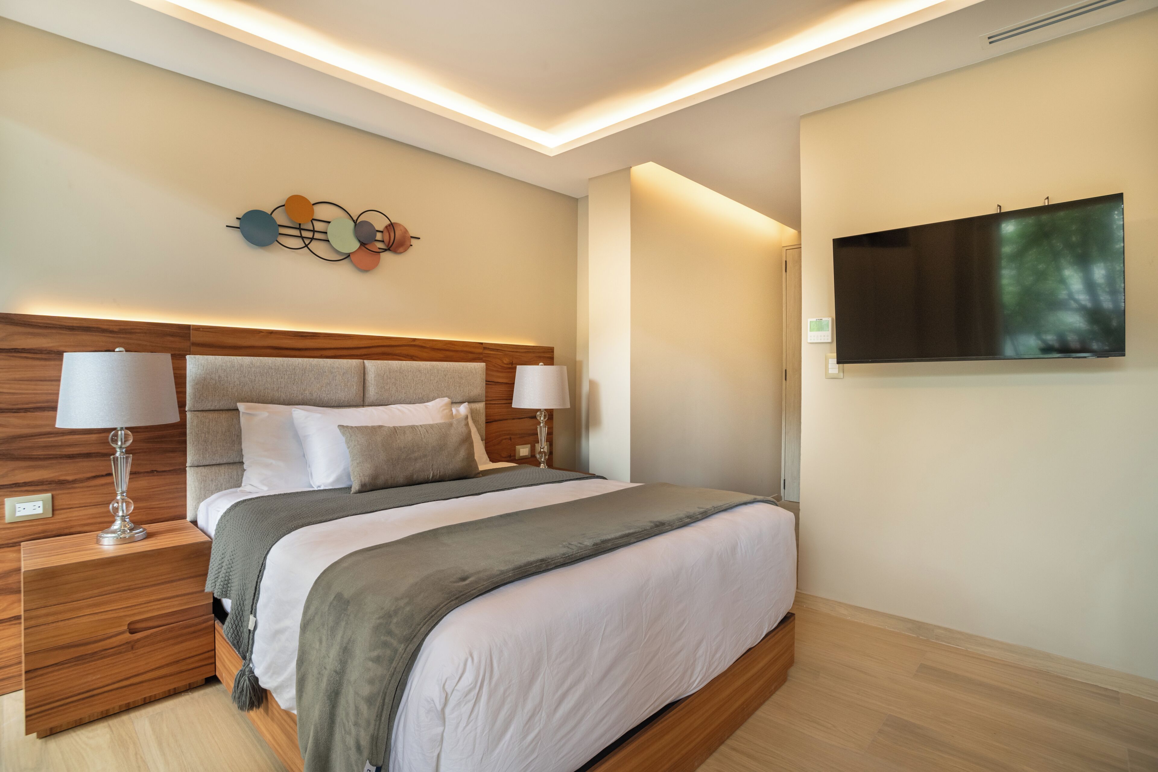 A modern hotel room with a wooden headboard, a flat-screen TV, a bedside table with a lamp, and a decorative wall piece.