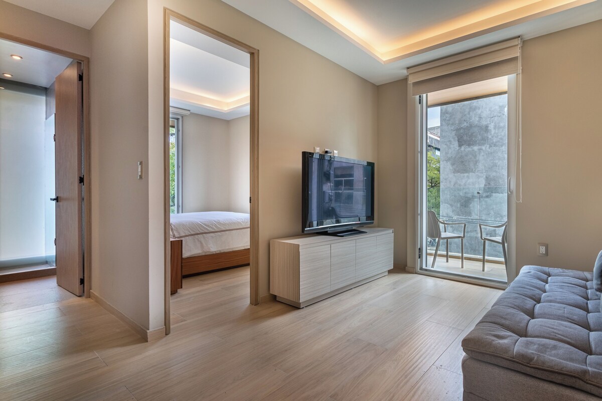 A modern bedroom with a bed, a TV on a cabinet, and a balcony with chairs.