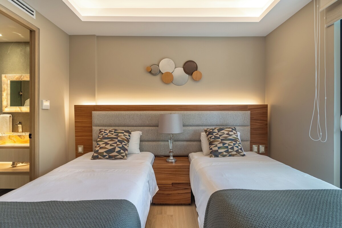 A hotel room with two beds, a wooden headboard, a wall-mounted lamp, and a mirror.