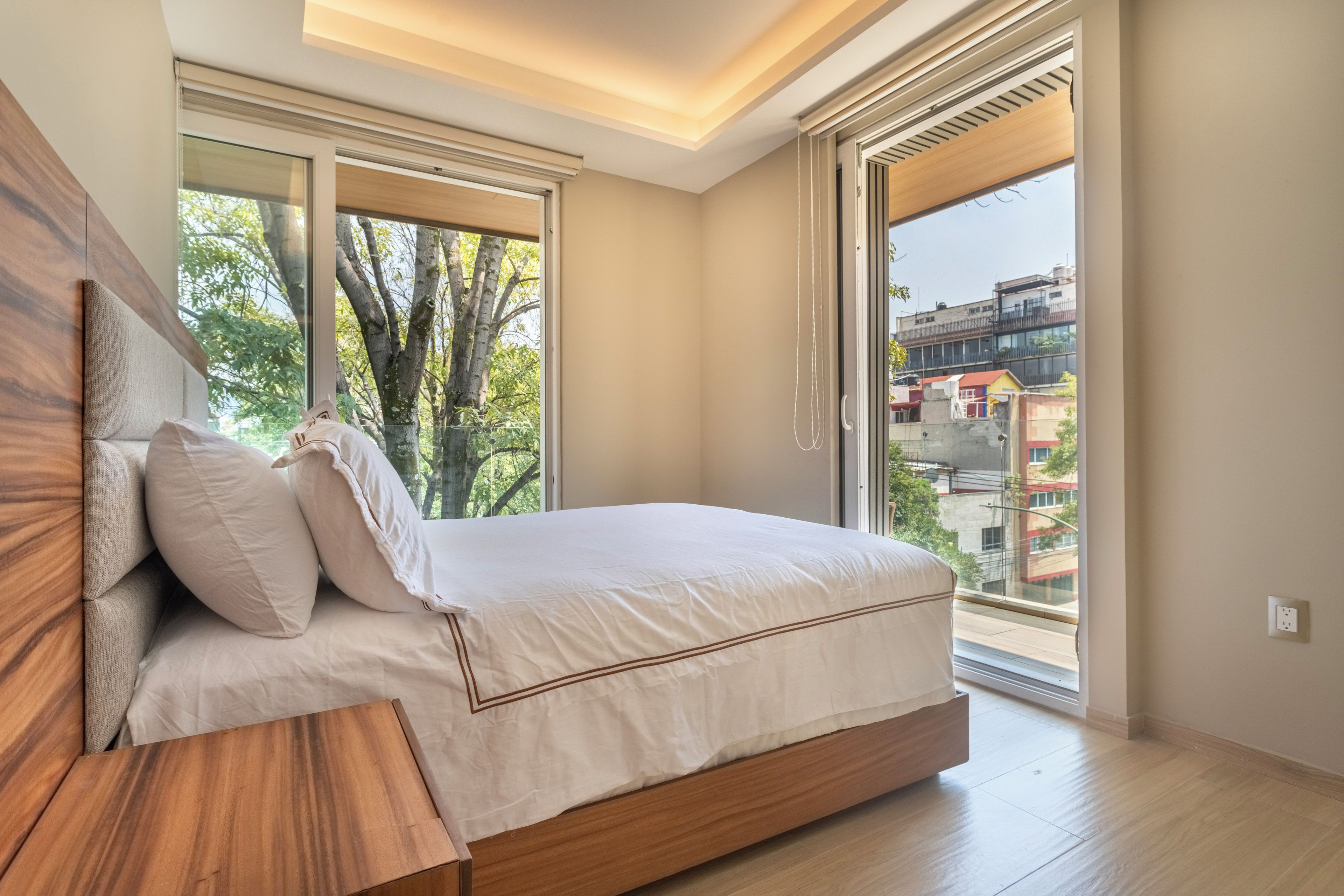 A bedroom with a bed, a wooden headboard, and a large window with a view of buildings and trees.