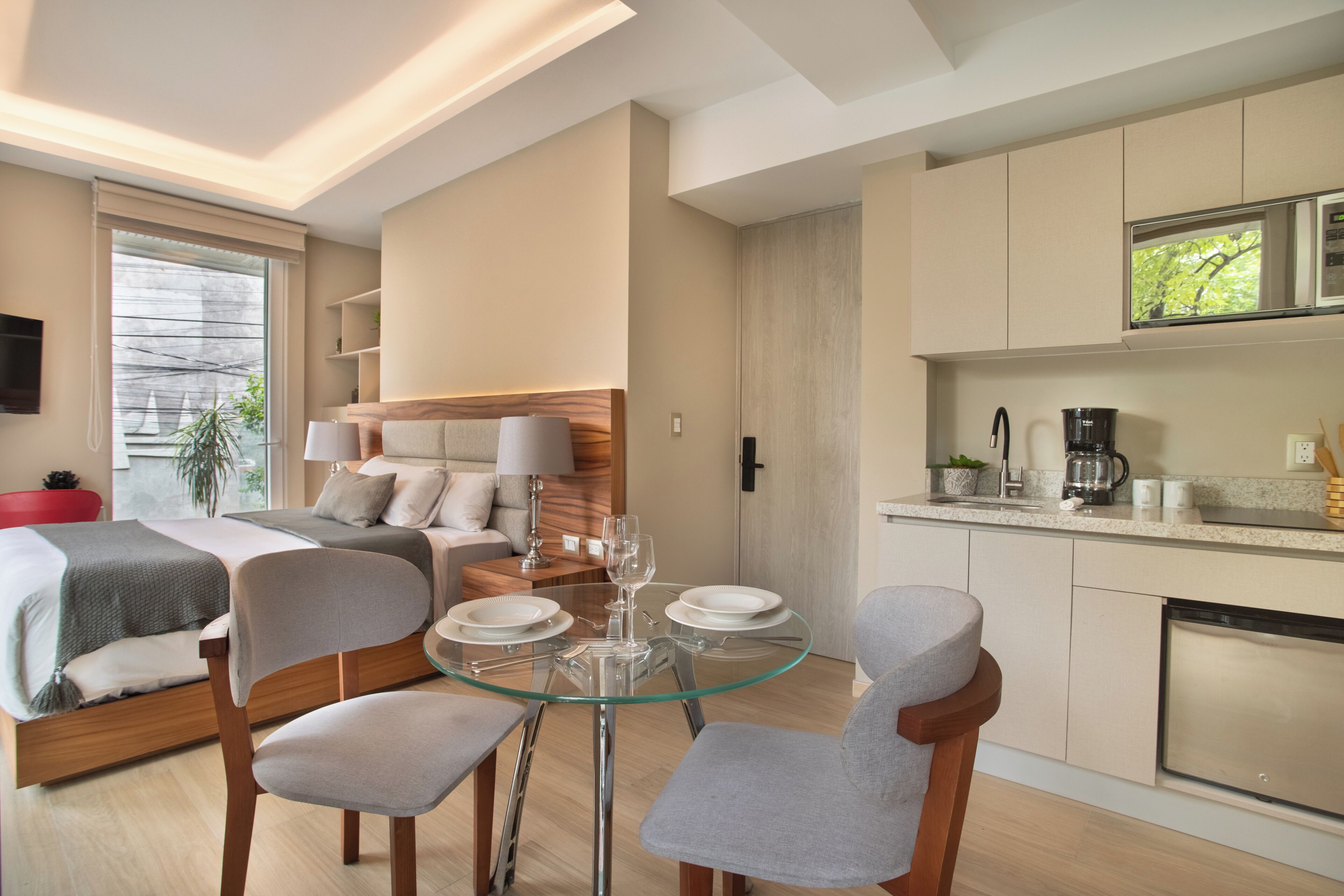 A modern hotel room with a bed, a glass dining table, and a kitchenette.