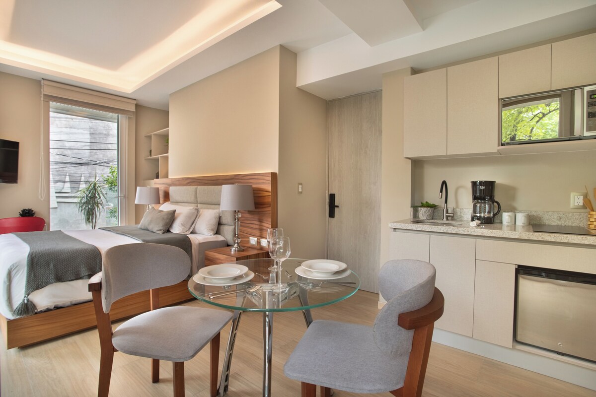 A modern hotel room with a bed, a glass dining table, and a kitchenette.