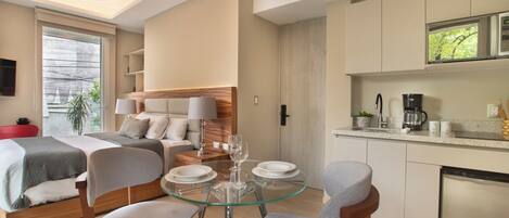 A modern hotel room with a bed, a glass dining table, and a kitchenette.