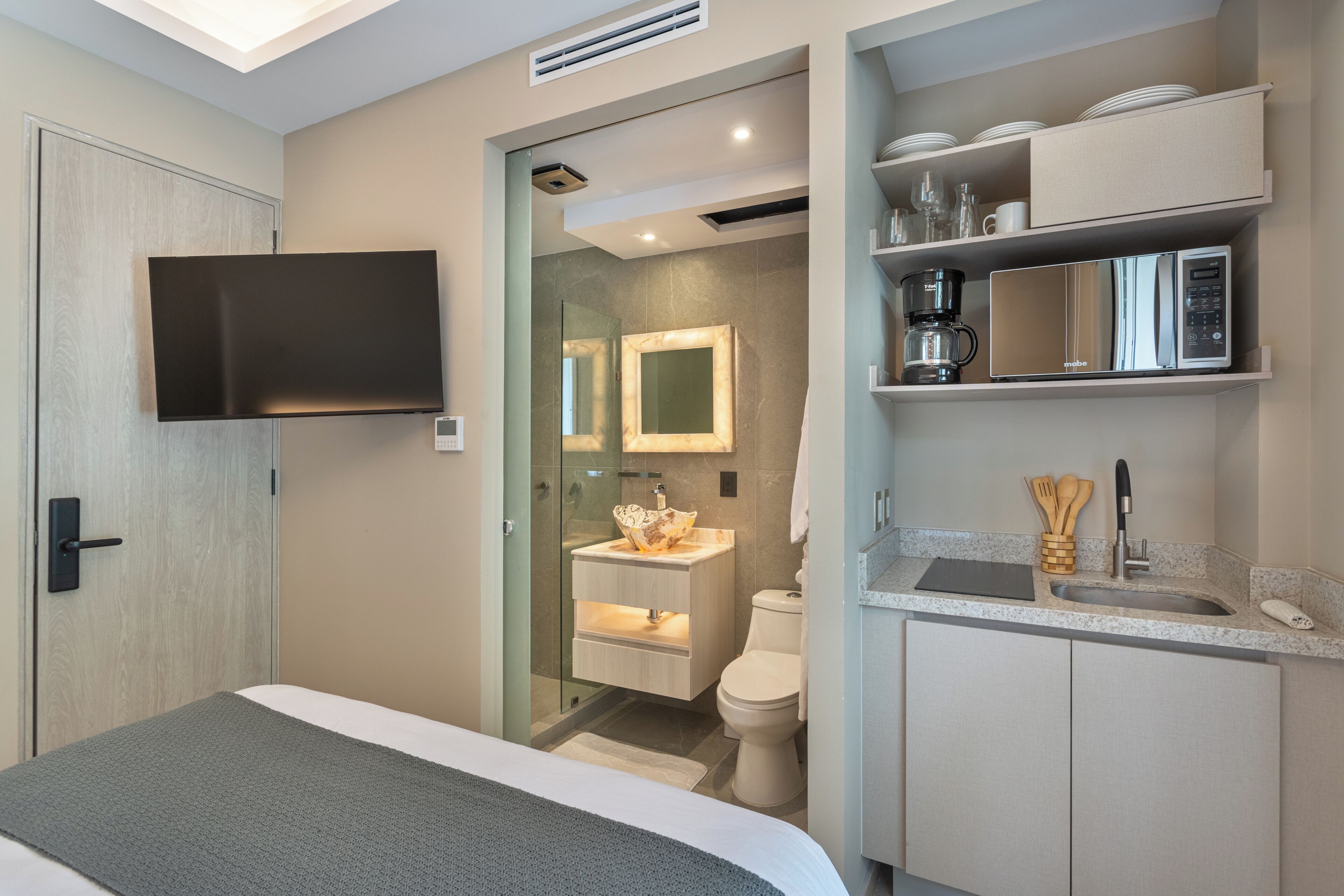 Habitación de hotel moderna con cama, televisor, baño con lavamanos e inodoro, y un área de cocina con microondas.
