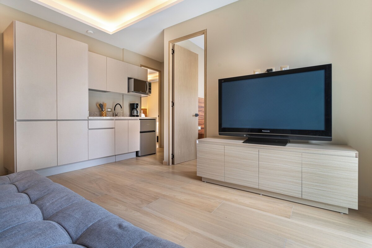 A modern living room with a flat-screen TV on a wooden cabinet, a grey sofa, and a kitchen area with cabinets and appliances.