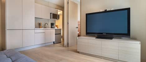 En moderne stue med et fladskærms-TV på et træskab, en grå sofa og et køkkenområde med skabe og hårde hvidevarer.