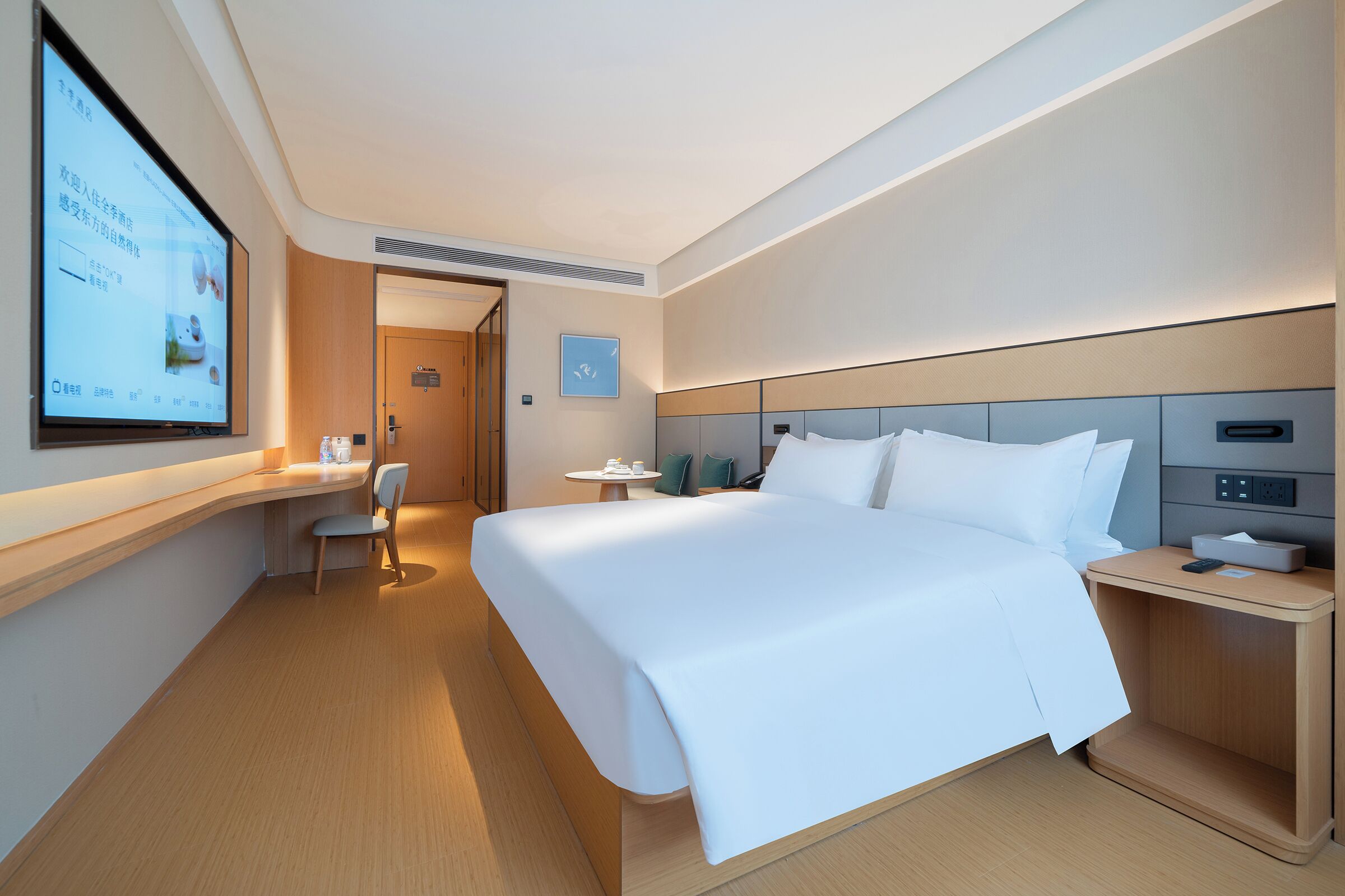 A hotel room with a large bed, a desk, a chair, a small table, and a TV screen.
