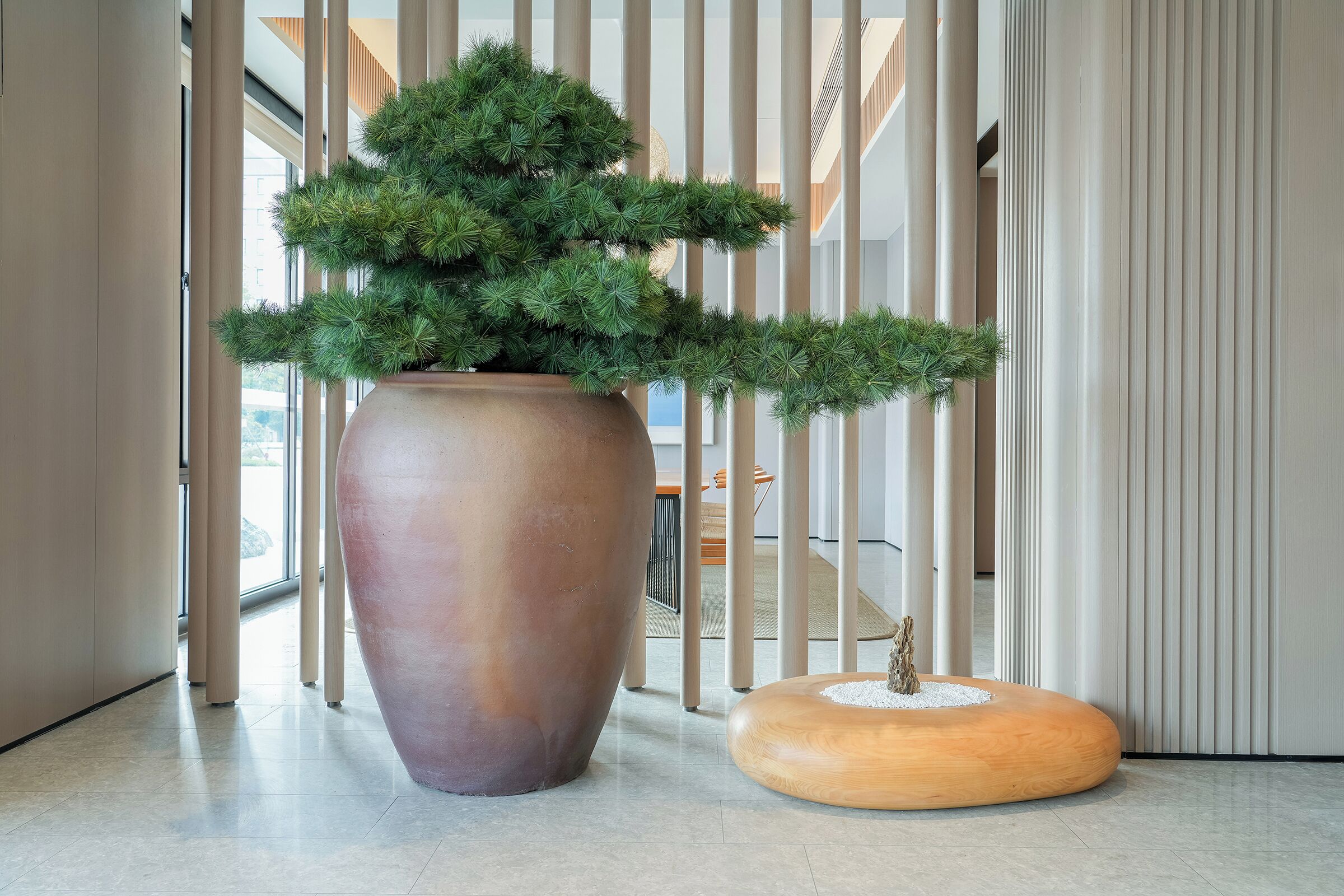 A large potted plant, a wooden stool, and a modern interior with vertical blinds.