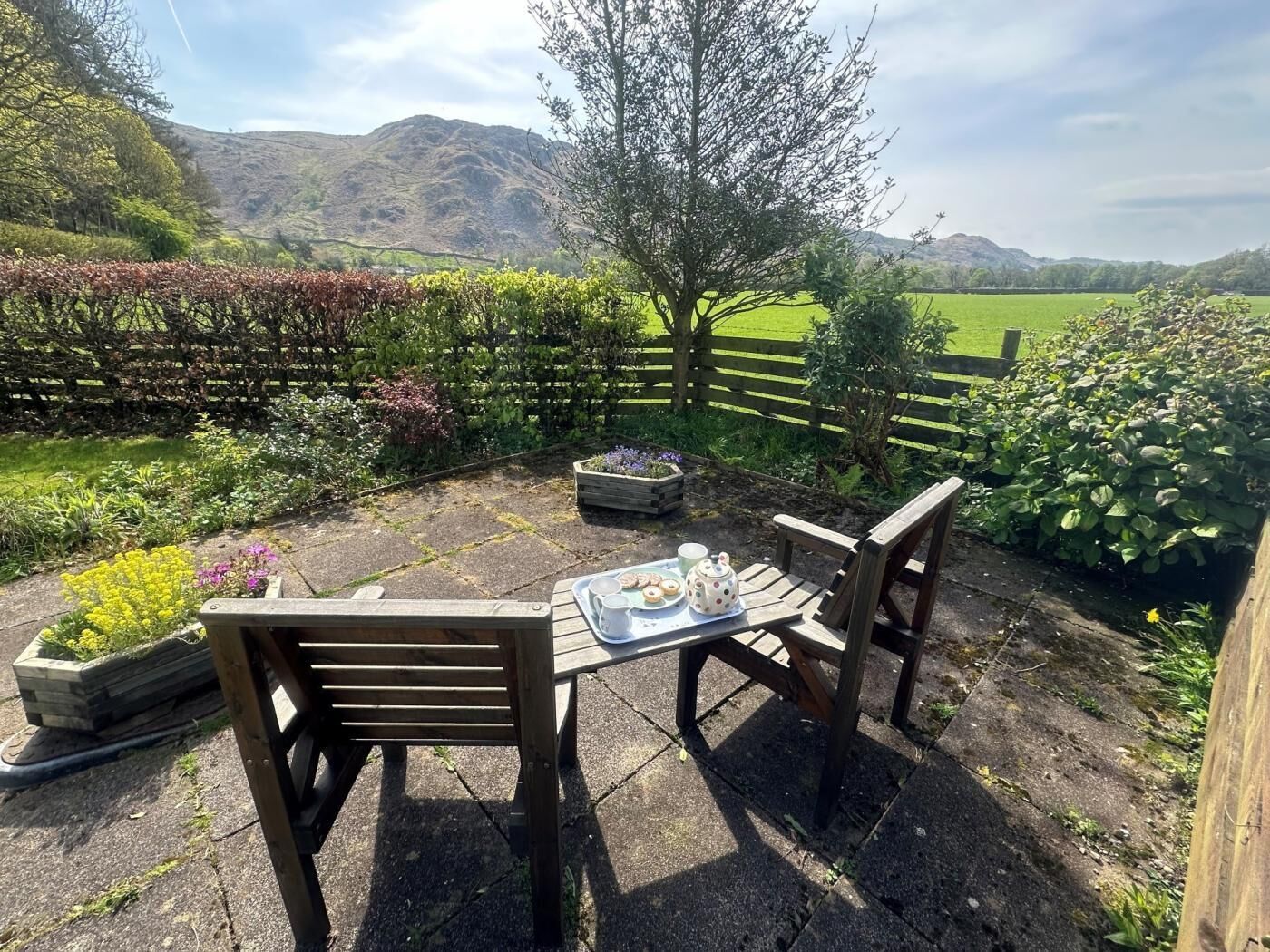 An outdoor seating area with a table set for tea, surrounded by a wooden fence and a view of a green field and hills.