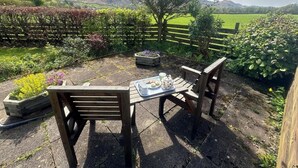 Un espace extérieur aménagé avec une table dressée pour le thé, entouré d’une clôture en bois et offrant une vue sur un champ verdoyant et des collines.