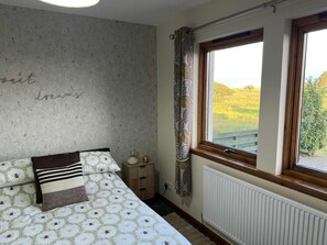 Un dormitorio con cama, mesita de noche, ventana con vistas a un campo y una pared con una frase escrita.