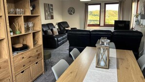 A living room with a sofa set, a wooden cabinet with glassware, a dining table, and a view of the outdoors through large windows. - 3 Bed in Newton Stewart (oc-87541) (Mochrum)