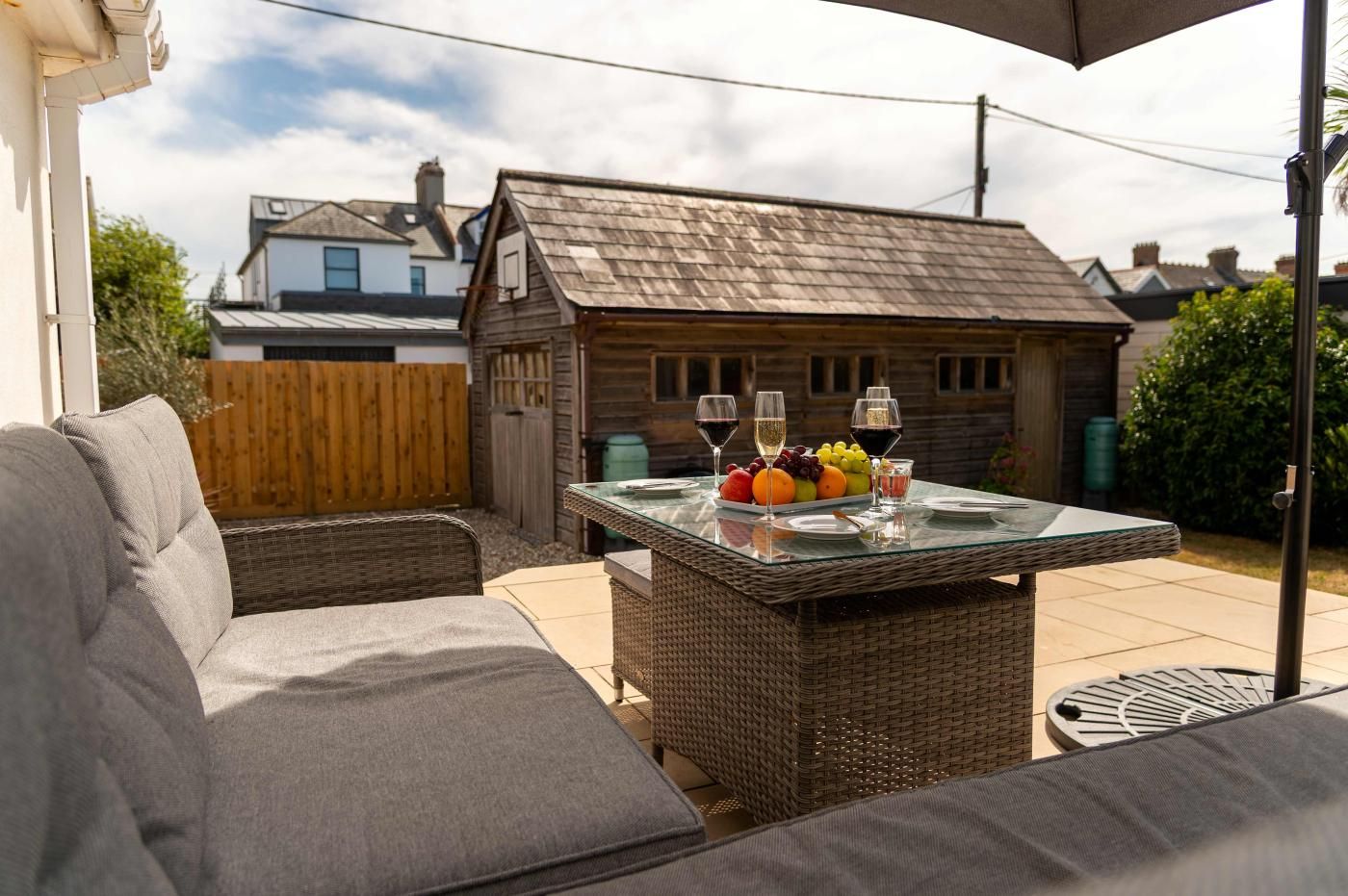 En udendørs terrasse med fletmøbler, et bord med glasplade, frugt og vinglas, samt en træbygning i baggrunden.