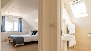 Quarto com cama, janela com cortinas, radiador e banheiro com pia e suporte para toalhas.