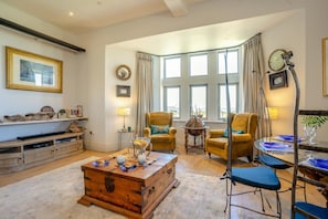 Ein Wohnzimmer mit Couchtisch aus Holz, zwei Sesseln, einer Anrichte und einem großen Fenster mit Vorhängen.