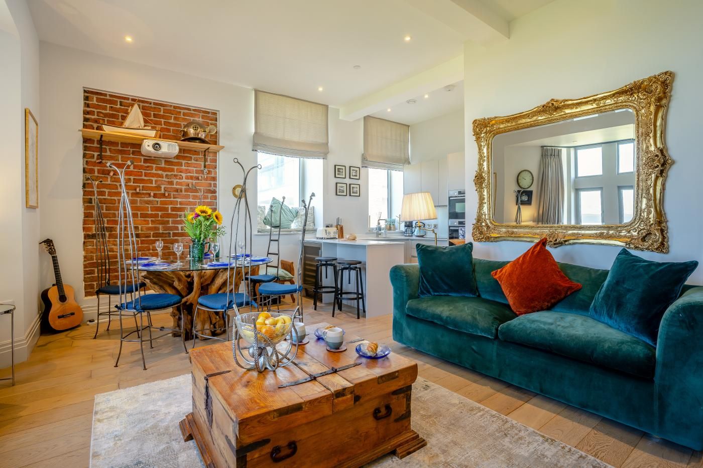 A living room with a green sofa, a wooden coffee table, and a brick wall. There is a dining area with blue chairs and a table. A large mirror with a gold frame is on the wall.