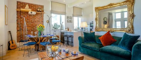A living room with a green sofa, a wooden coffee table, and a brick wall. There is a dining area with blue chairs and a table. A large mirror with a gold frame is on the wall.