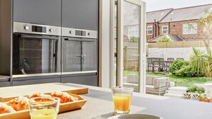 Eine moderne Küche mit zwei Backöfen, ein Essbereich mit Tisch und Stühlen sowie Blick auf einen Garten mit Zaun und Grünflächen.