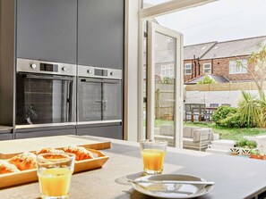 A modern kitchen with two ovens, a dining area with a table and chairs, and a view of a backyard with a fence and greenery.