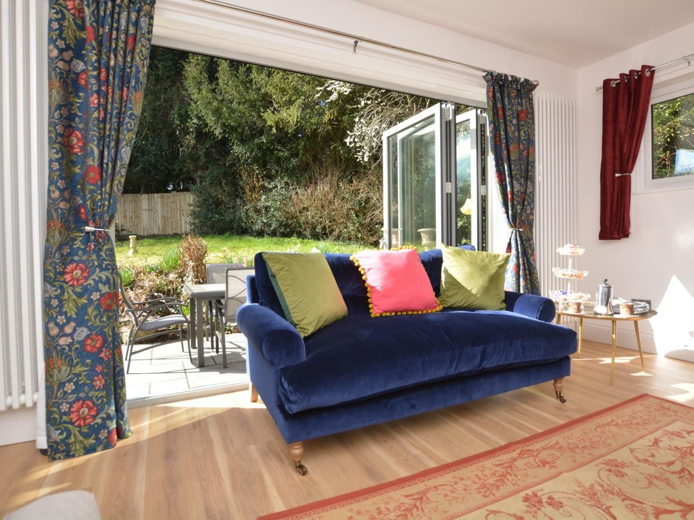 A blue sofa with colorful cushions in a room with a large window, a small table with a tray, and a patterned rug.
