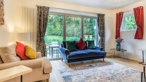 A living room with a sofa, a blue armchair, a coffee table, and a rug. There are two windows with curtains, a chandelier, and a small table with a vase. - 4 Bed in Bakewell (oc-87419) (Bakewell)