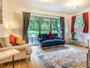 Ein Wohnzimmer mit einer Couch, einem blauen Sessel, einem Couchtisch und einem Teppich. Zwei Fenster mit Vorhängen, eine Kristallleuchter und ein kleiner Tisch mit einer Vase sind ebenfalls vorhanden.